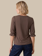 A woman shown from the back wearing a brown three-quarter sleeve top with ruffled cuffs and dark blue jeans, set against a solid beige background.
