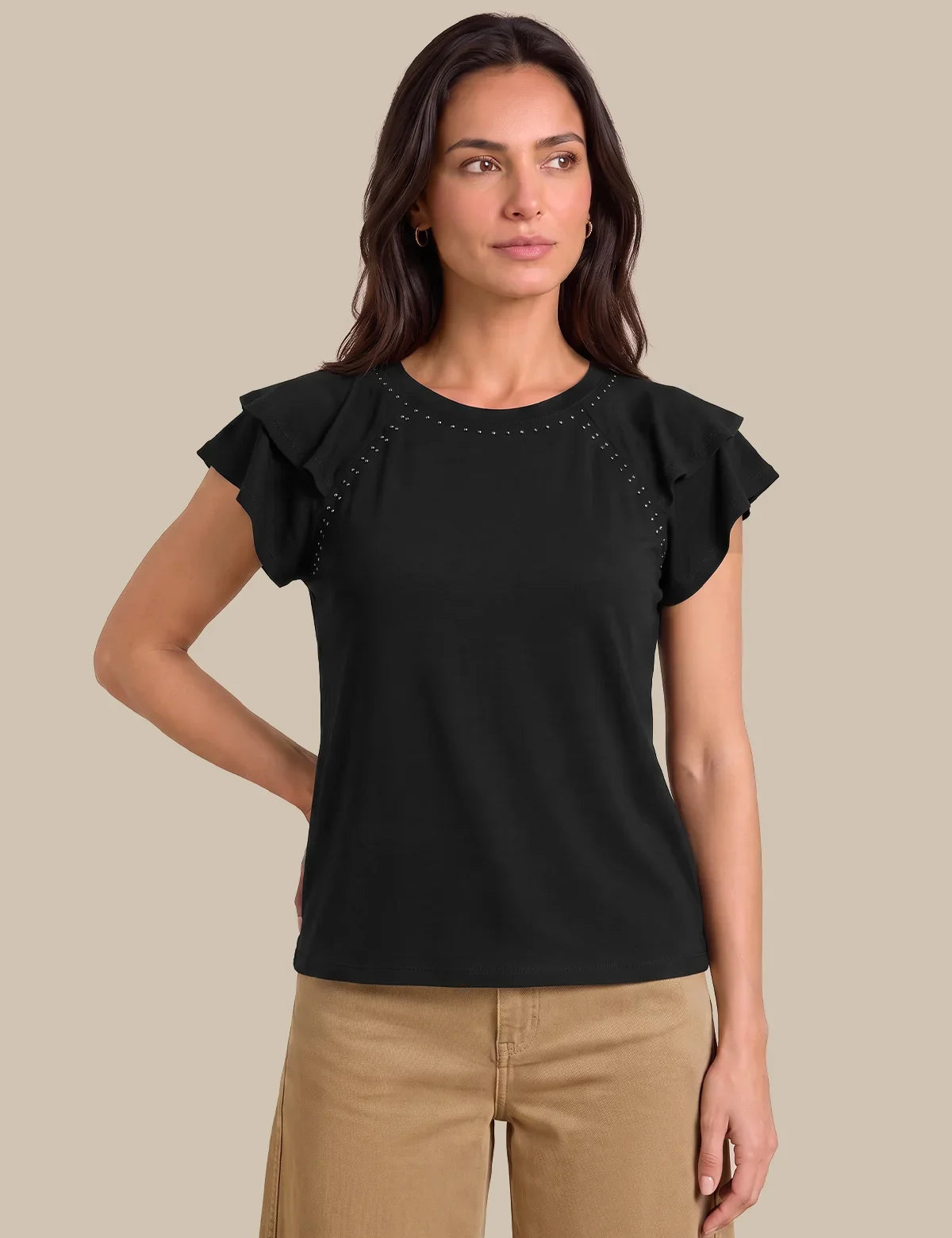 A woman modeling a black short-sleeve top with ruffled sleeves and decorative stud detailing along the shoulder seams, paired with tan pants, against a solid beige background.