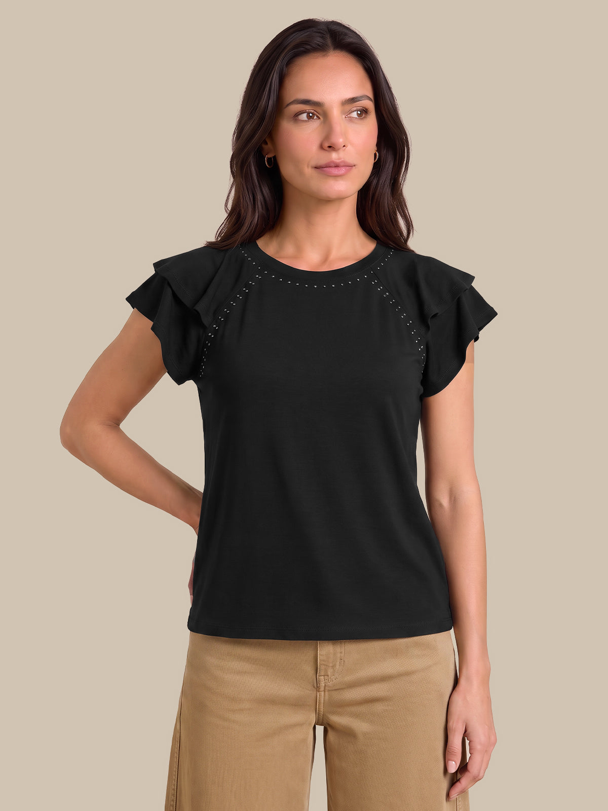 A woman modeling a black short-sleeve top with ruffled sleeves and decorative stud detailing along the shoulder seams, paired with tan pants, against a solid beige background.