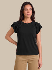 A woman modeling a black short-sleeve top with ruffled sleeves and decorative stud detailing along the shoulder seams, paired with tan pants, against a solid beige background.