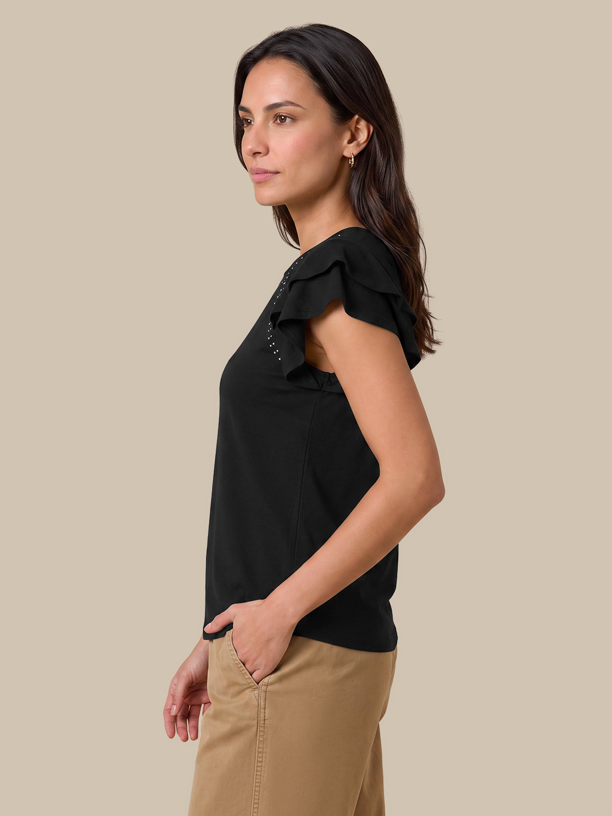 A woman shown in side profile wearing a black ruffled short-sleeve top and tan pants against a plain beige studio background.