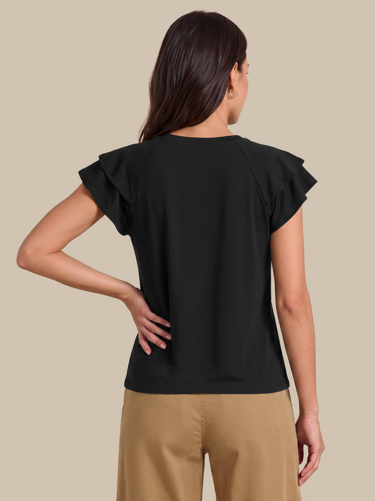 A woman shown from the back wearing a black short-sleeve top with ruffled edges and tan pants, against a plain beige studio background.