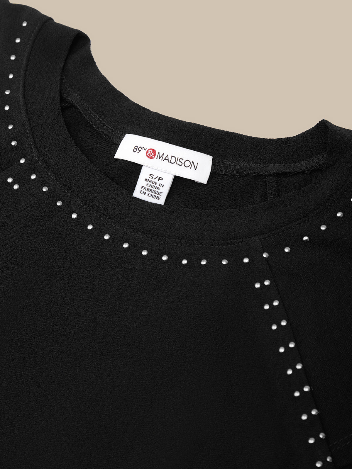 Close-up of a black crewneck top with silver stud embellishments around the neckline, showing a white brand label that reads 69& MADISON.