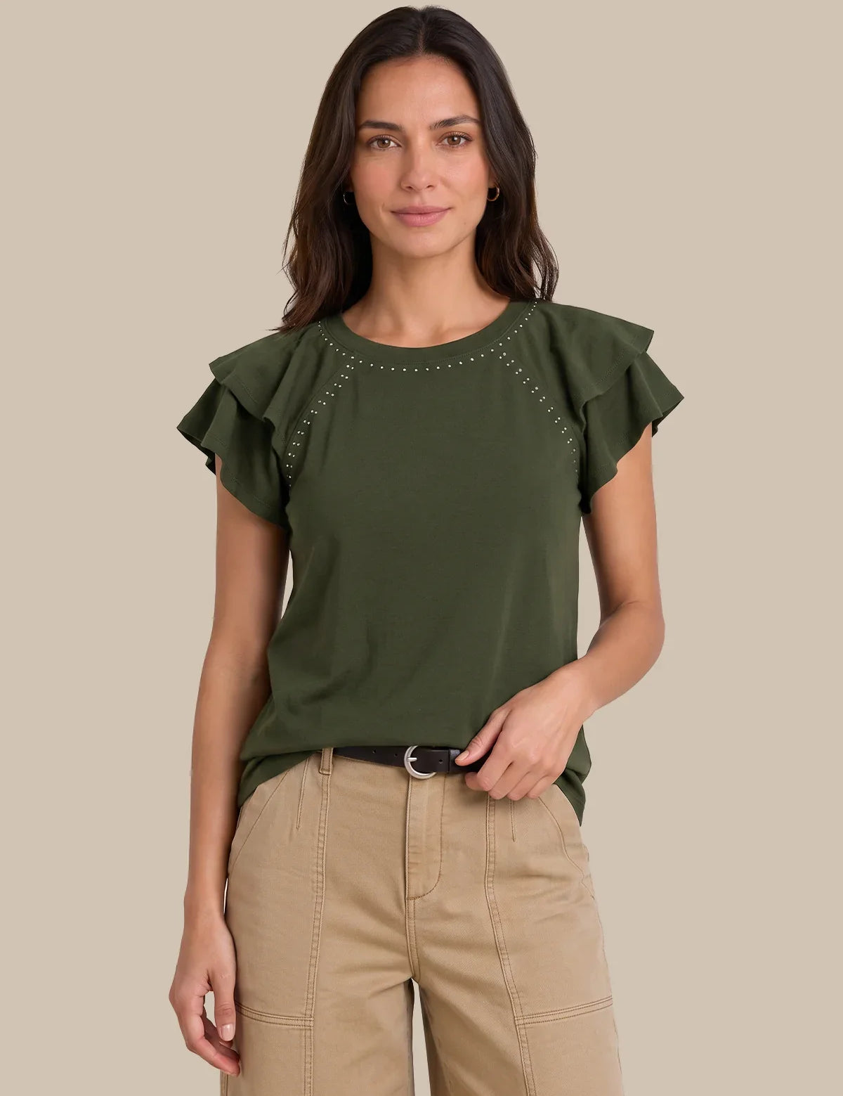A woman modeling an olive green short-sleeve top with ruffled sleeves and silver stud detailing around the neckline, paired with beige pants, against a solid beige background.
