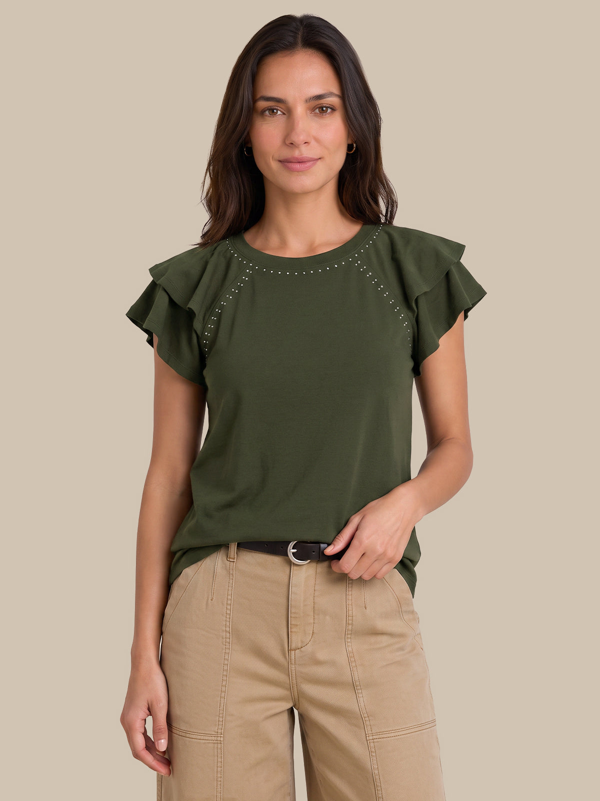 A woman modeling an olive green short-sleeve top with ruffled sleeves and silver stud detailing around the neckline, paired with beige pants, against a solid beige background.