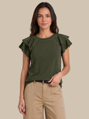 A woman modeling an olive green short-sleeve top with ruffled sleeves and silver stud detailing around the neckline, paired with beige pants, against a solid beige background.