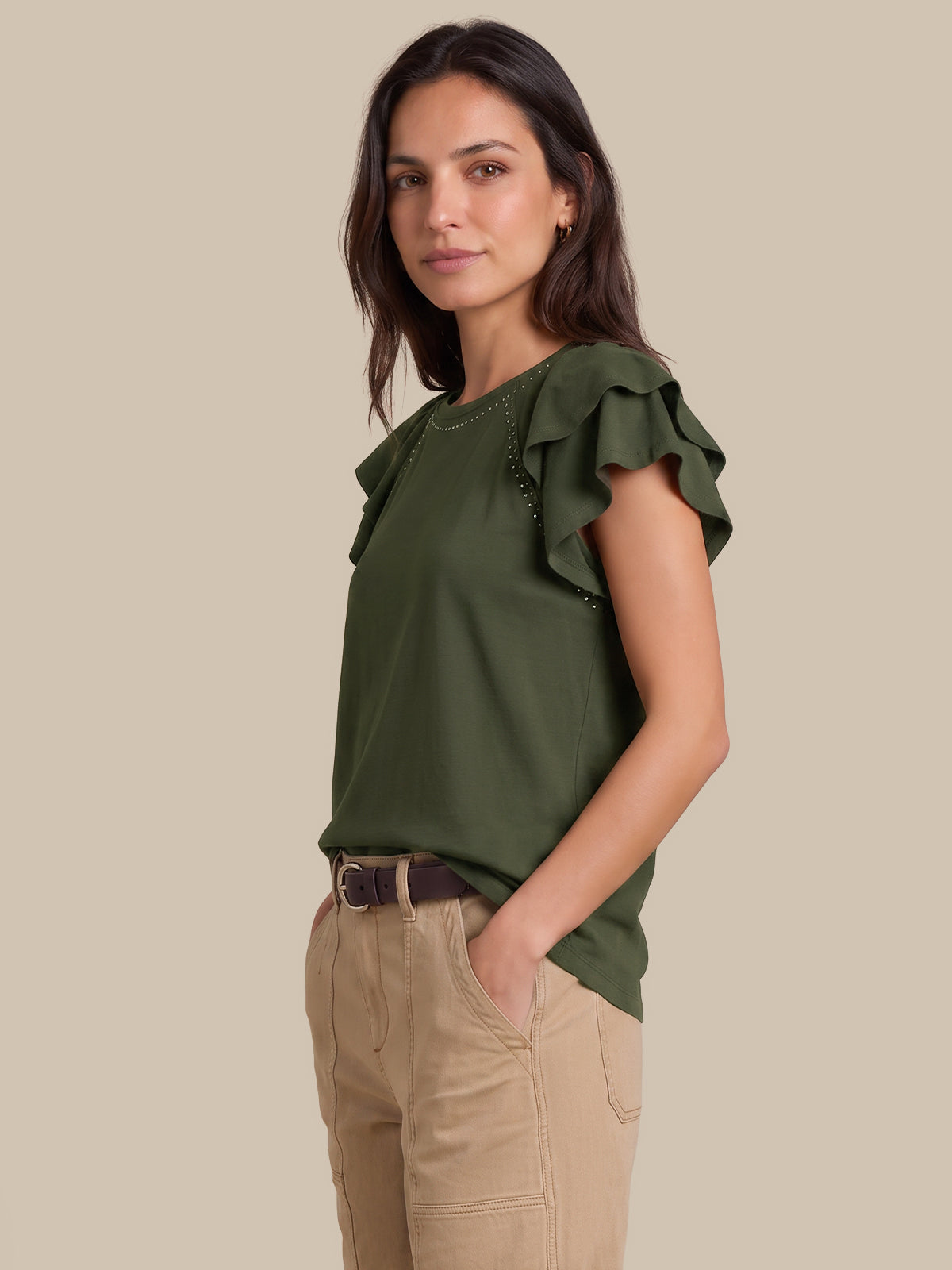 A woman modeling an olive green ruffled short-sleeve top with beaded trim, paired with beige pants, against a solid beige background.