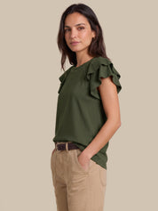 A woman modeling an olive green ruffled short-sleeve top with beaded trim, paired with beige pants, against a solid beige background.