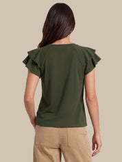 A woman shown from the back wearing an olive green short-sleeve top with ruffled shoulder details and beige pants, set against a solid beige background.