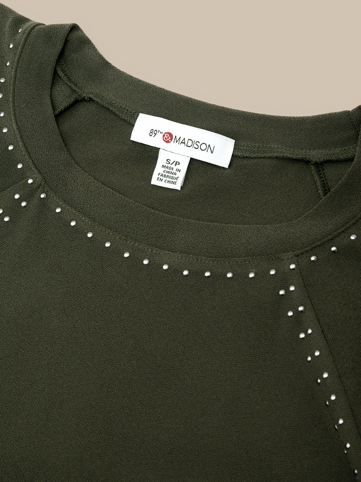 Close-up of a dark olive green top with silver stud embellishments around the neckline and shoulder, featuring a visible '69 MADISON' neck label.