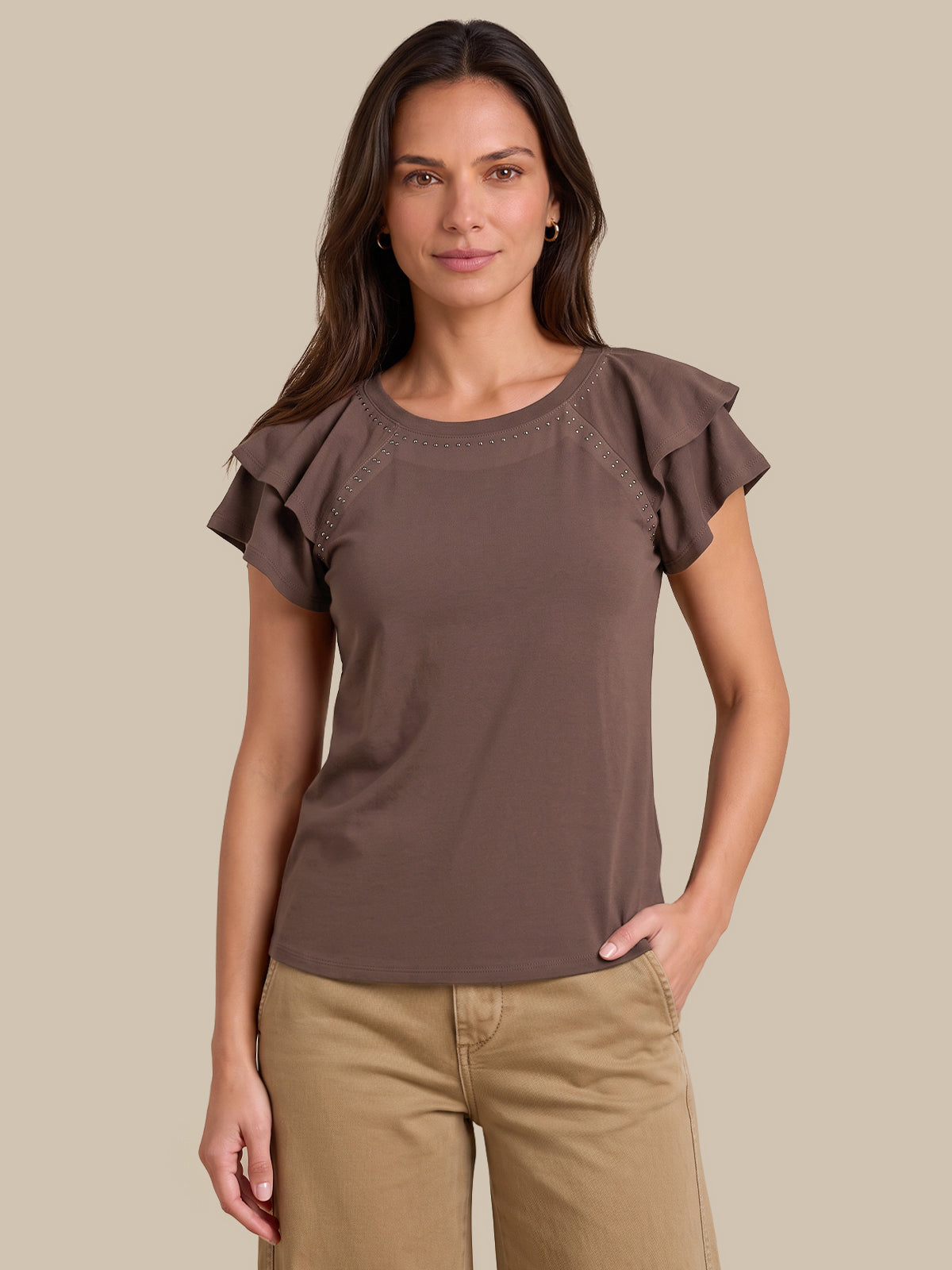 A woman modeling a brown short-sleeve top with ruffled cap sleeves and a crew neckline, paired with tan pants, against a solid beige background.
