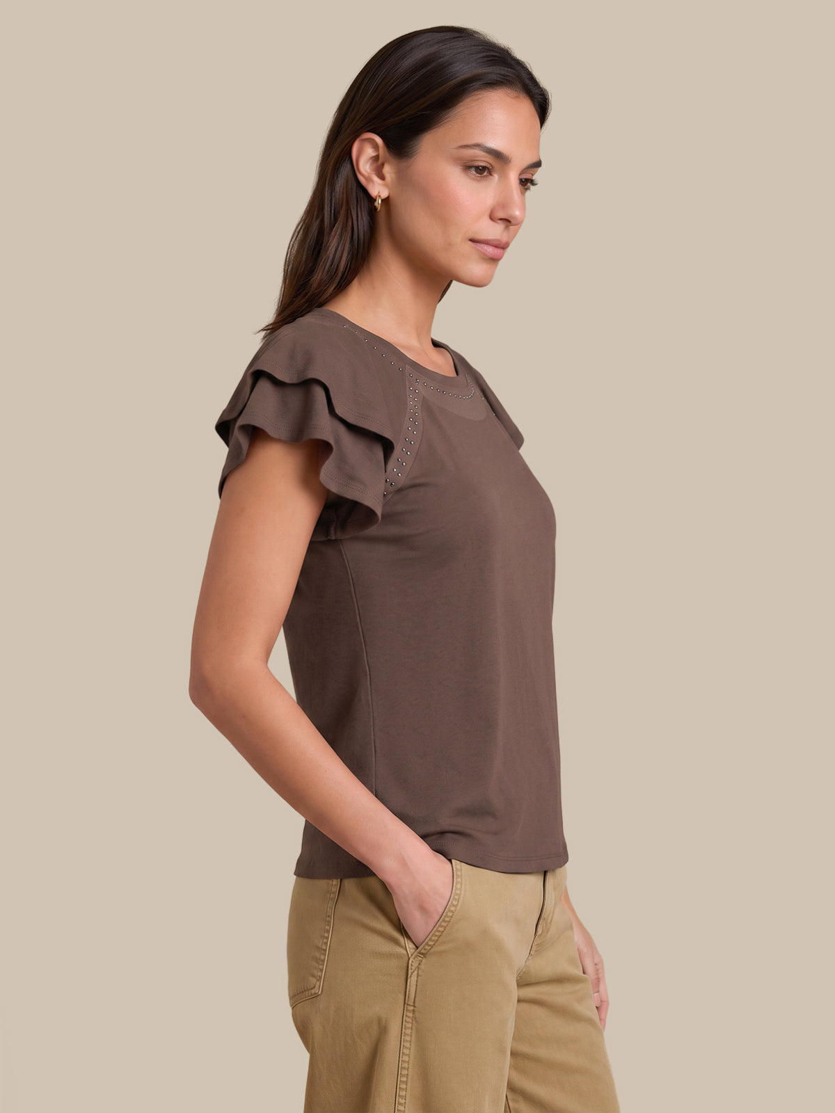 A woman wearing a brown ruffled short-sleeve top and khaki pants, shown in profile against a solid beige background.