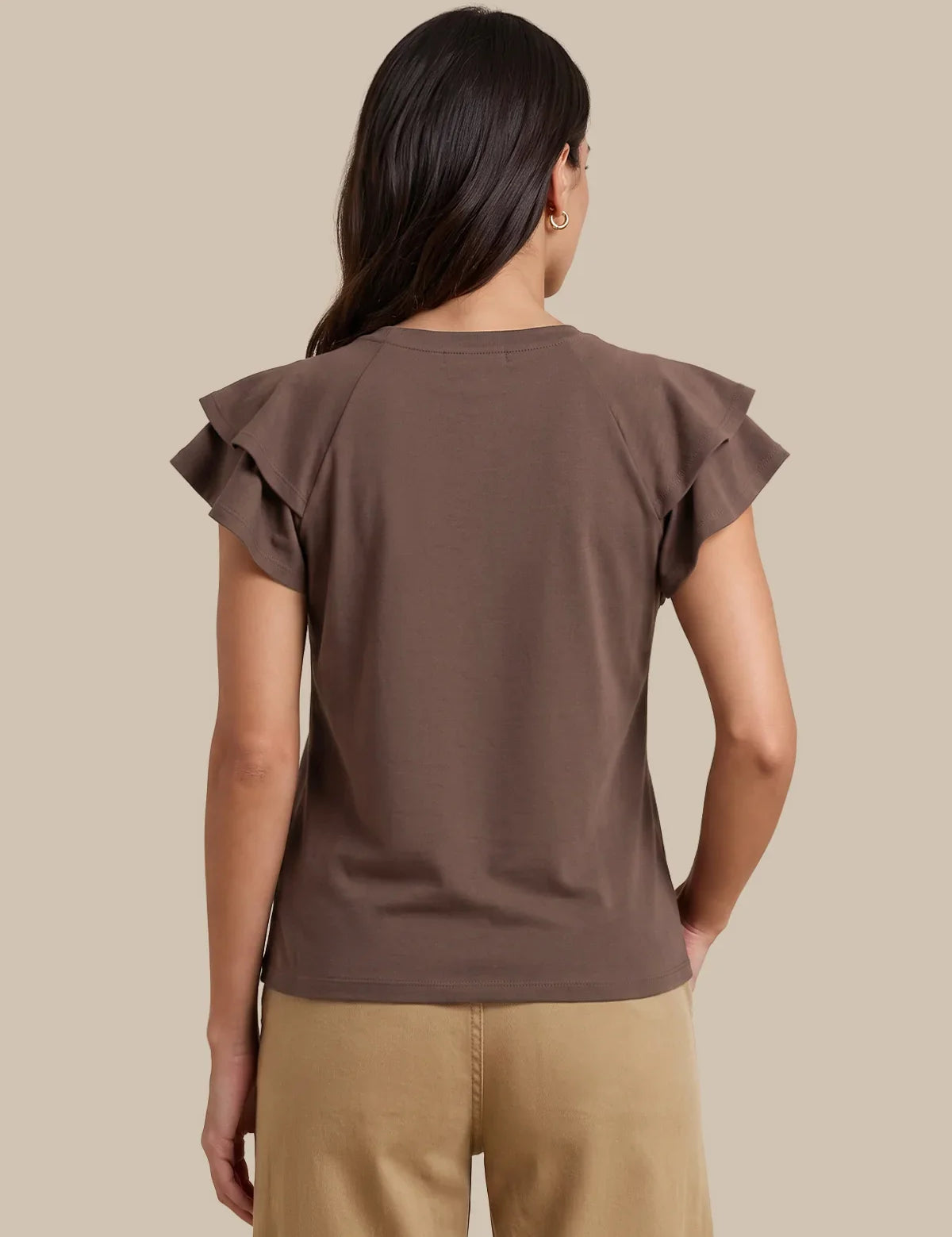A woman viewed from the back wearing a brown short-sleeve top with ruffled shoulder details and tan pants, set against a solid beige background.