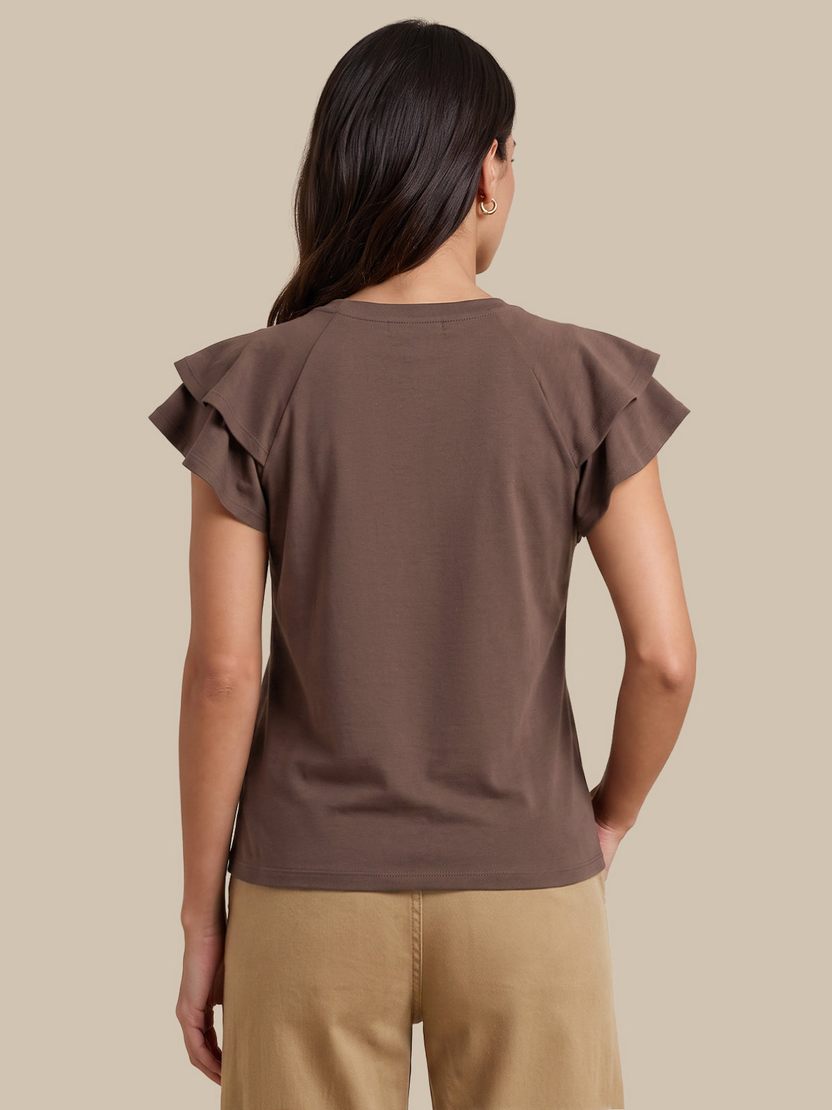 A woman viewed from the back wearing a brown short-sleeve top with ruffled shoulder details and tan pants, set against a solid beige background.