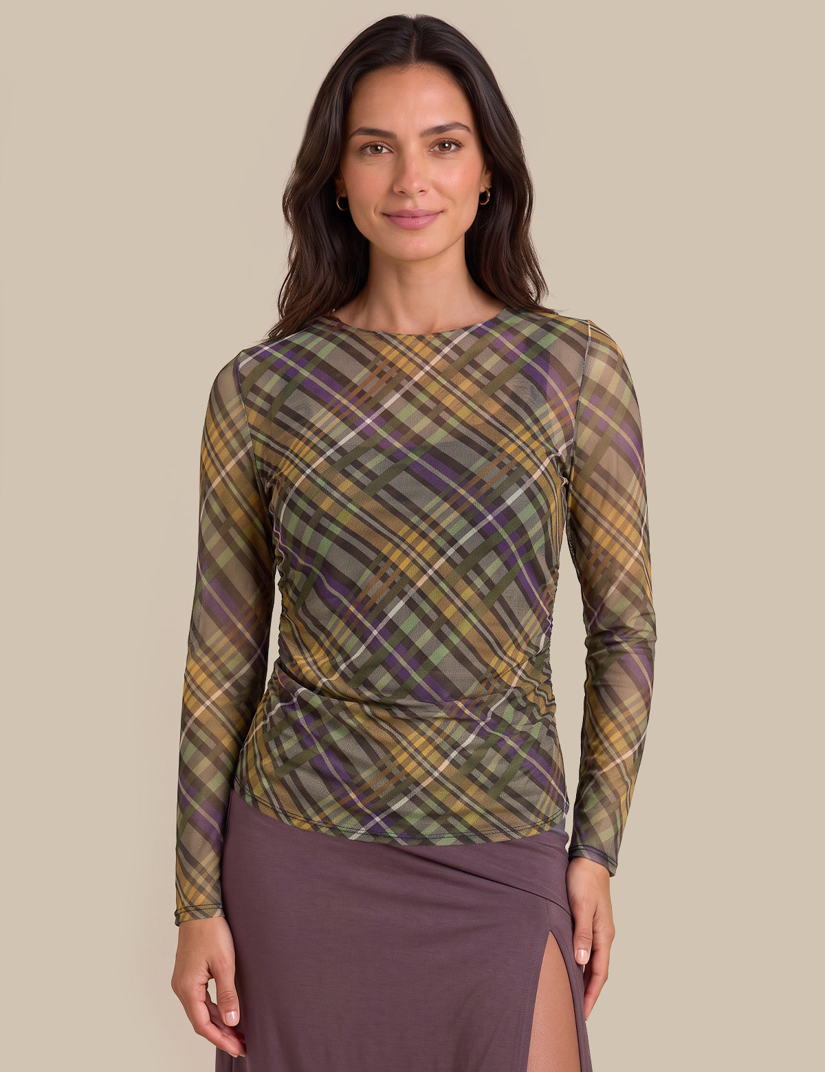 A woman modeling a long-sleeve knit top with a diagonal plaid pattern in olive green, mustard yellow, and purple, paired with a mauve skirt, against a solid beige background.