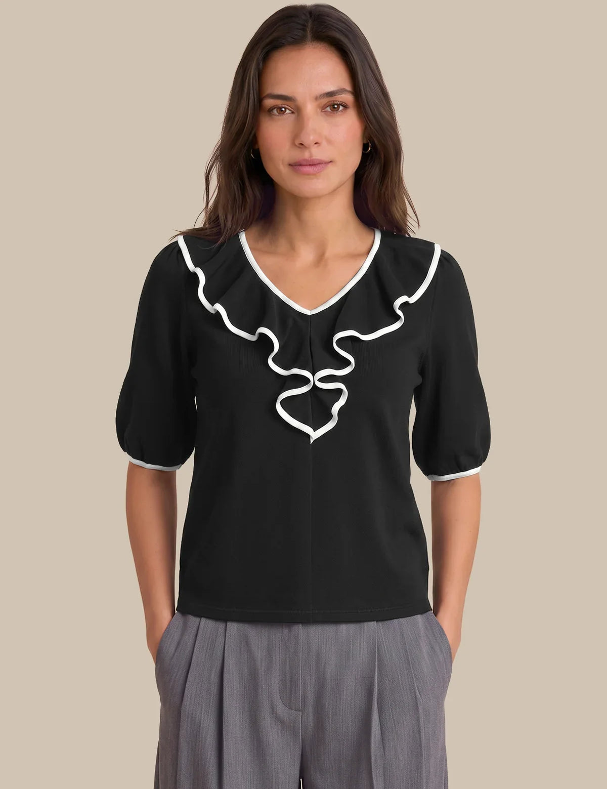 A woman modeling a black short-sleeve blouse with white ruffle trim and a white bow detail at the V-neck, paired with gray pleated trousers, against a solid beige background.