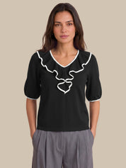 A woman modeling a black short-sleeve blouse with white ruffle trim and a white bow detail at the V-neck, paired with gray pleated trousers, against a solid beige background.