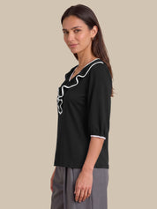 A woman modeling a black short-sleeve blouse with white ruffle trim along the neckline and cuffs, paired with gray trousers, against a solid beige background.