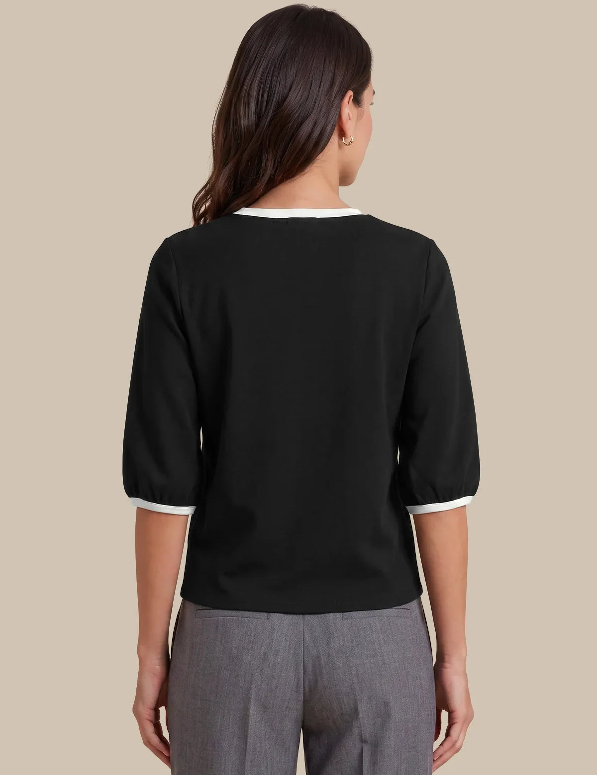 A woman shown from the back wearing a black three-quarter sleeve top with white trim at the collar and cuffs, paired with gray trousers, against a solid beige background.