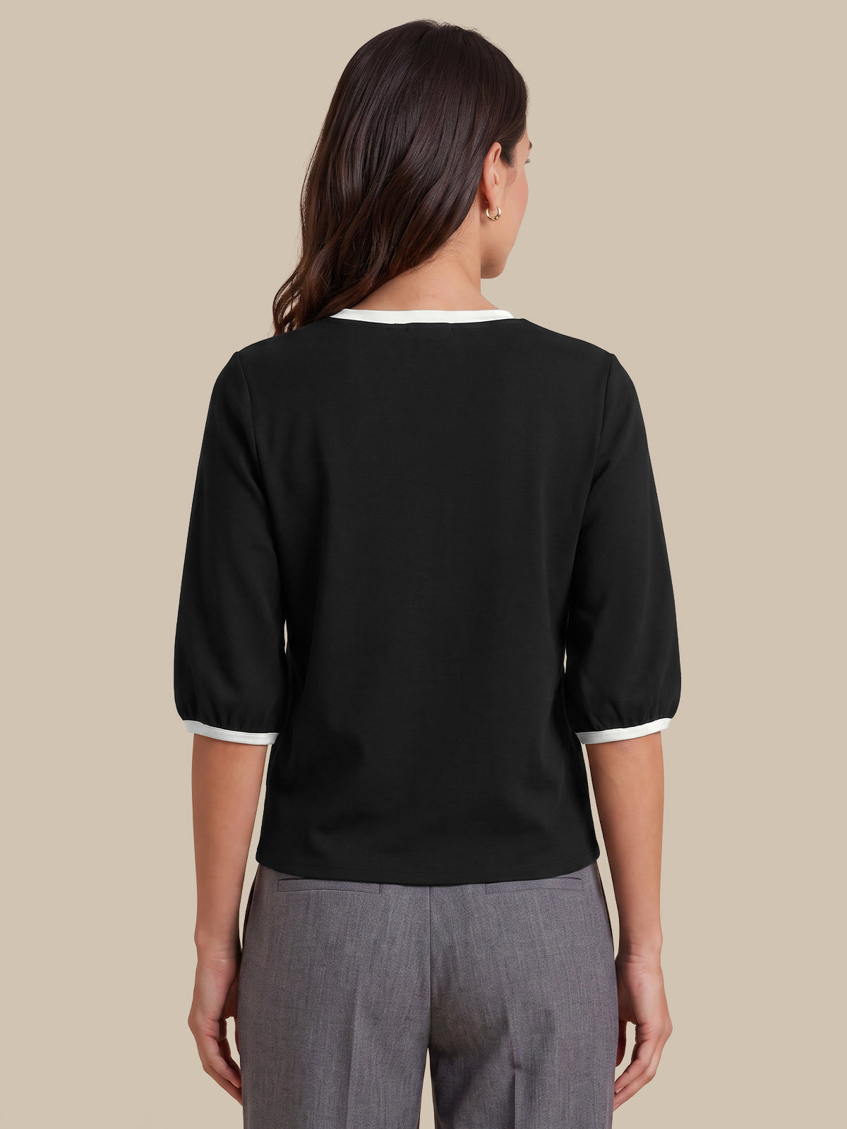 A woman shown from the back wearing a black three-quarter sleeve top with white trim at the collar and cuffs, paired with gray trousers, against a solid beige background.