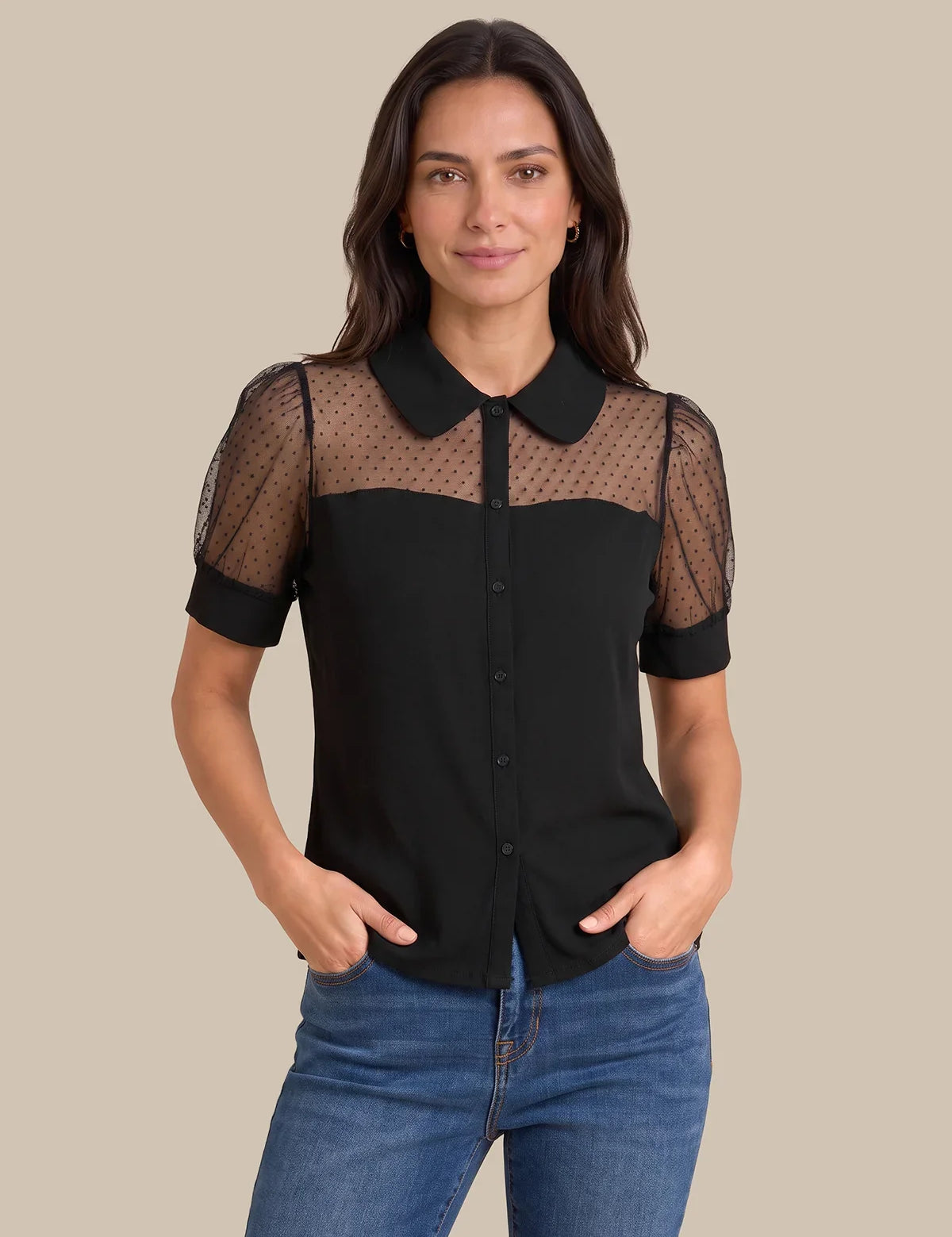 A woman modeling a black short-sleeve blouse with sheer mesh shoulder panels and a Peter Pan collar, paired with blue jeans against a solid beige background.