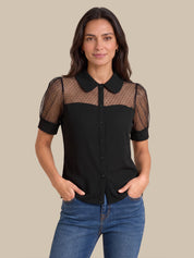 A woman modeling a black short-sleeve blouse with sheer mesh shoulder panels and a Peter Pan collar, paired with blue jeans against a solid beige background.