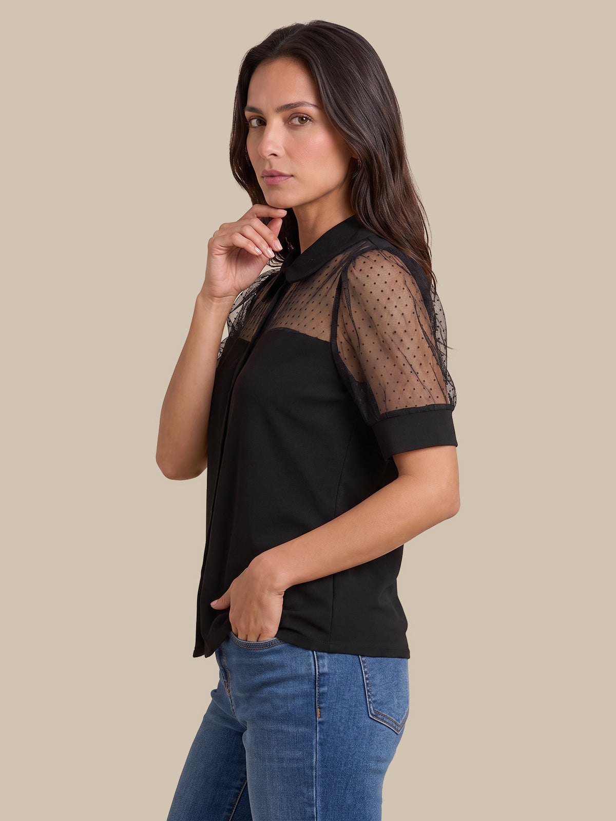 A woman modeling a black short-sleeve blouse with sheer polka-dot mesh sleeves, paired with blue jeans, against a solid beige background.