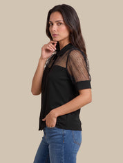 A woman modeling a black short-sleeve blouse with sheer polka-dot mesh sleeves, paired with blue jeans, against a solid beige background.