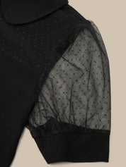 Close-up of a black short-sleeve top with sheer dotted mesh puff sleeves and a solid black collar, shown against a light beige background.