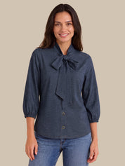 A woman wearing a dark slate blue long-sleeve blouse with a large bow at the collar and button-front closure, paired with blue jeans, against a solid beige background.