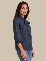 A woman wearing a dark navy blue blouse with a bow detail at the collar and three-quarter sleeves, paired with blue jeans, set against a solid beige background.