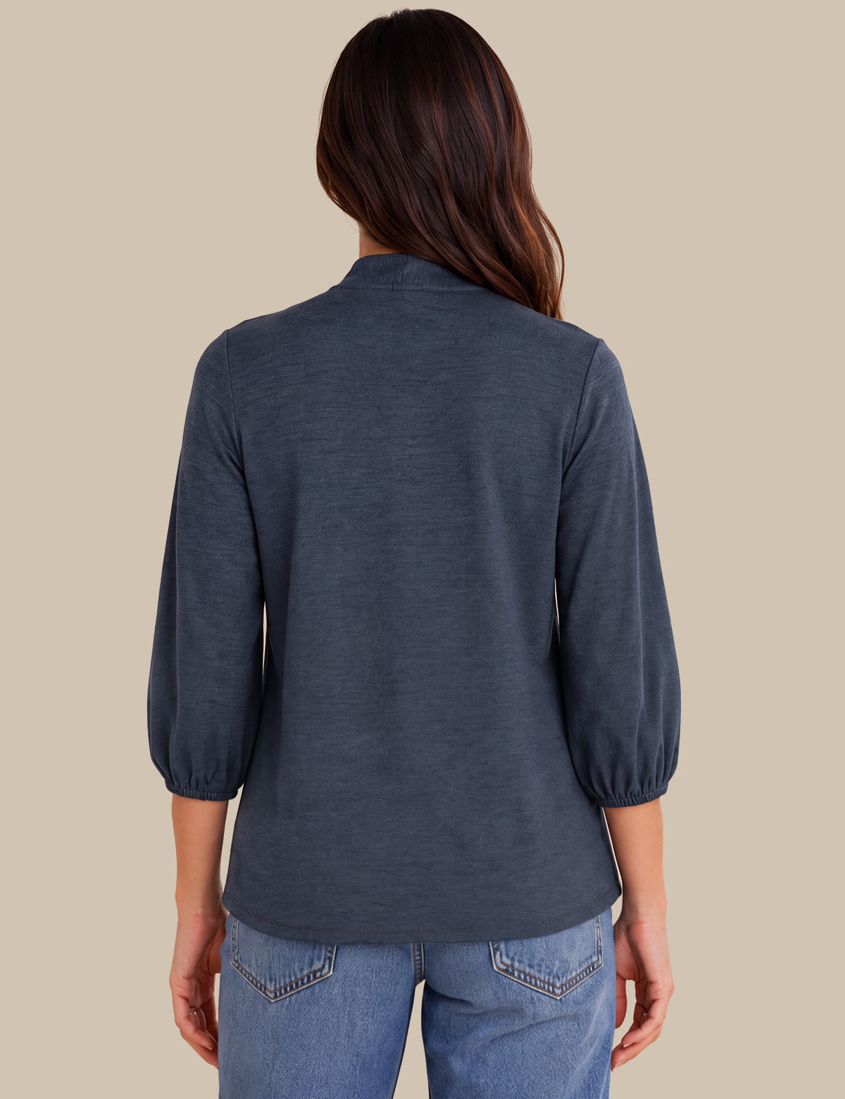 A woman viewed from the back wearing a slate blue three-quarter sleeve top with gathered cuffs and blue jeans, set against a solid beige background.