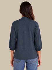 A woman viewed from the back wearing a slate blue three-quarter sleeve top with gathered cuffs and blue jeans, set against a solid beige background.