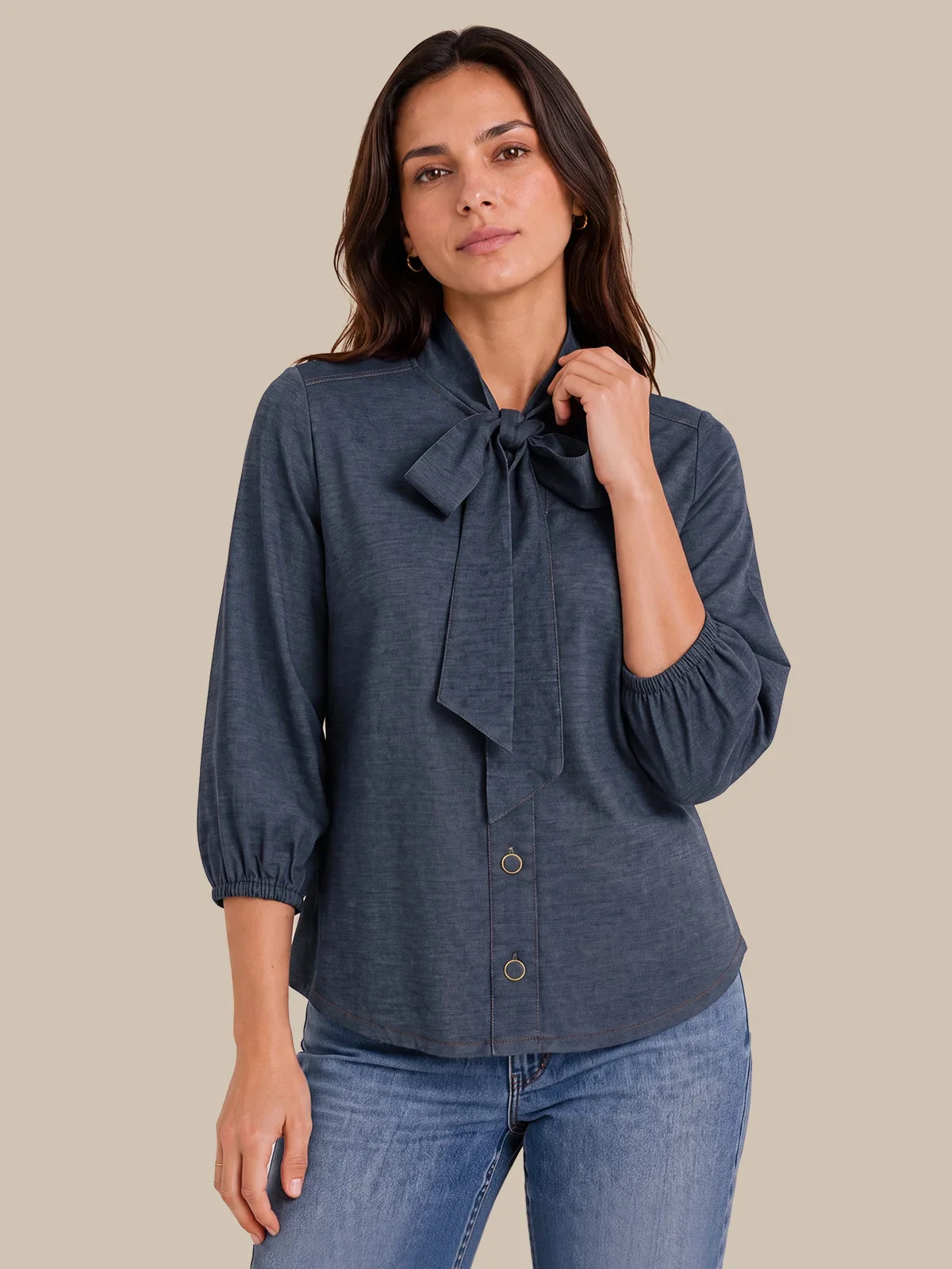 A woman modeling a slate blue long-sleeve blouse with a large bow at the collar and button-front closure, paired with blue jeans against a solid beige background.