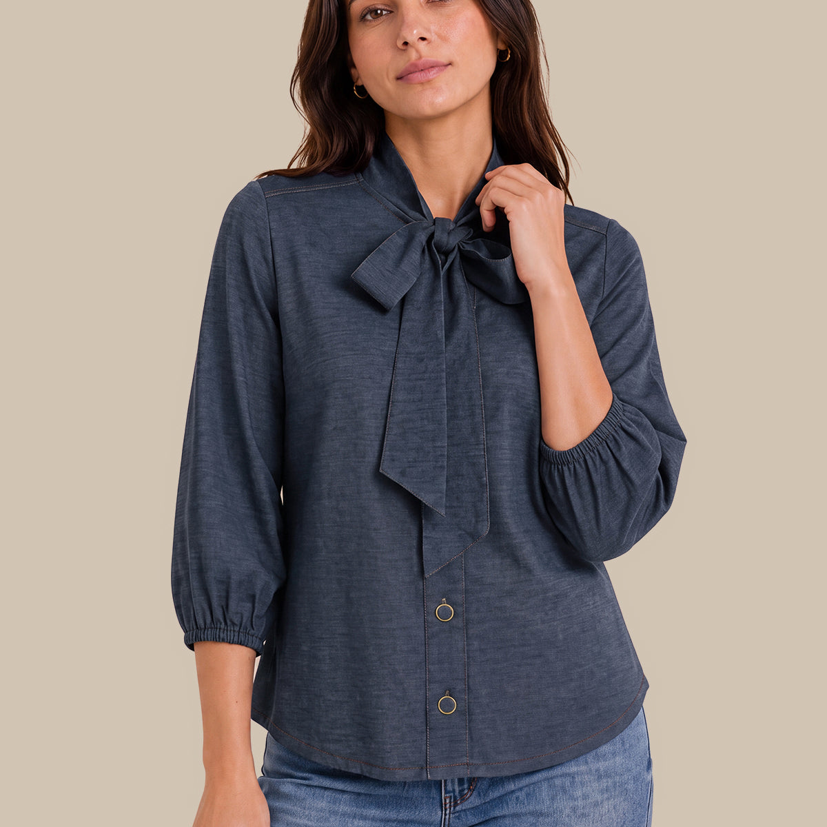 A woman modeling a dark slate blue long-sleeve blouse with a large bow at the collar and button-front closure, paired with blue jeans against a solid beige background.