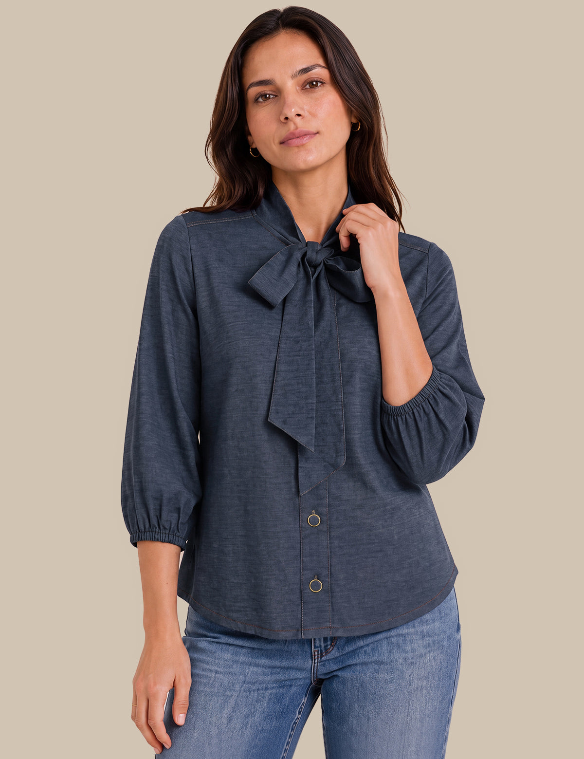 A woman modeling a dark slate blue long-sleeve blouse with a large bow at the collar and button-front closure, paired with blue jeans against a solid beige background.