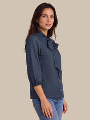 A woman wearing a dark navy blue blouse with a bow detail at the collar and three-quarter sleeves, paired with blue jeans, set against a solid beige background.