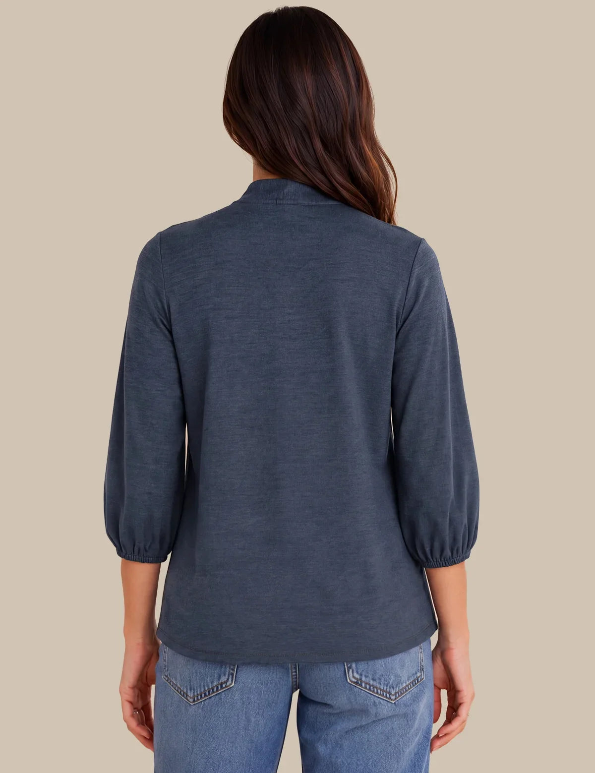 A woman viewed from the back wearing a slate blue three-quarter sleeve top and light blue jeans, set against a solid beige background.