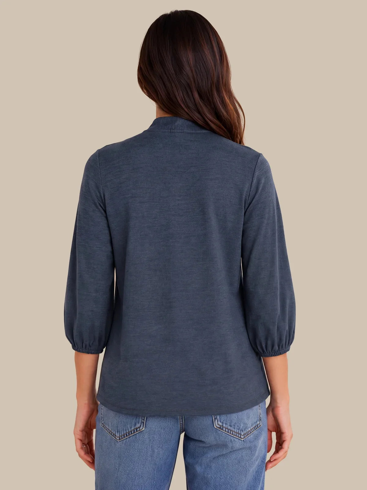 A woman viewed from the back wearing a slate blue three-quarter sleeve top and light blue jeans, set against a solid beige background.