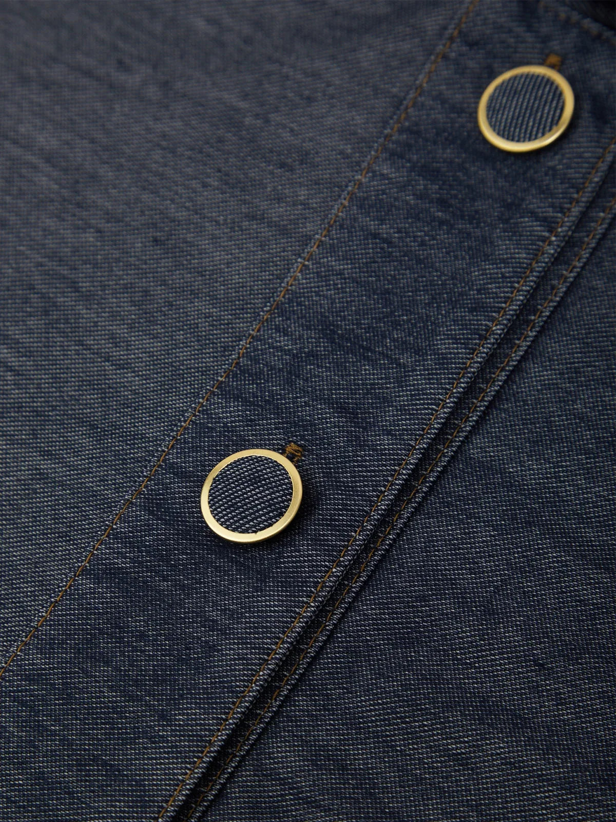 Close-up of dark indigo denim fabric with two round gold-tone metal snap buttons and contrast stitching.