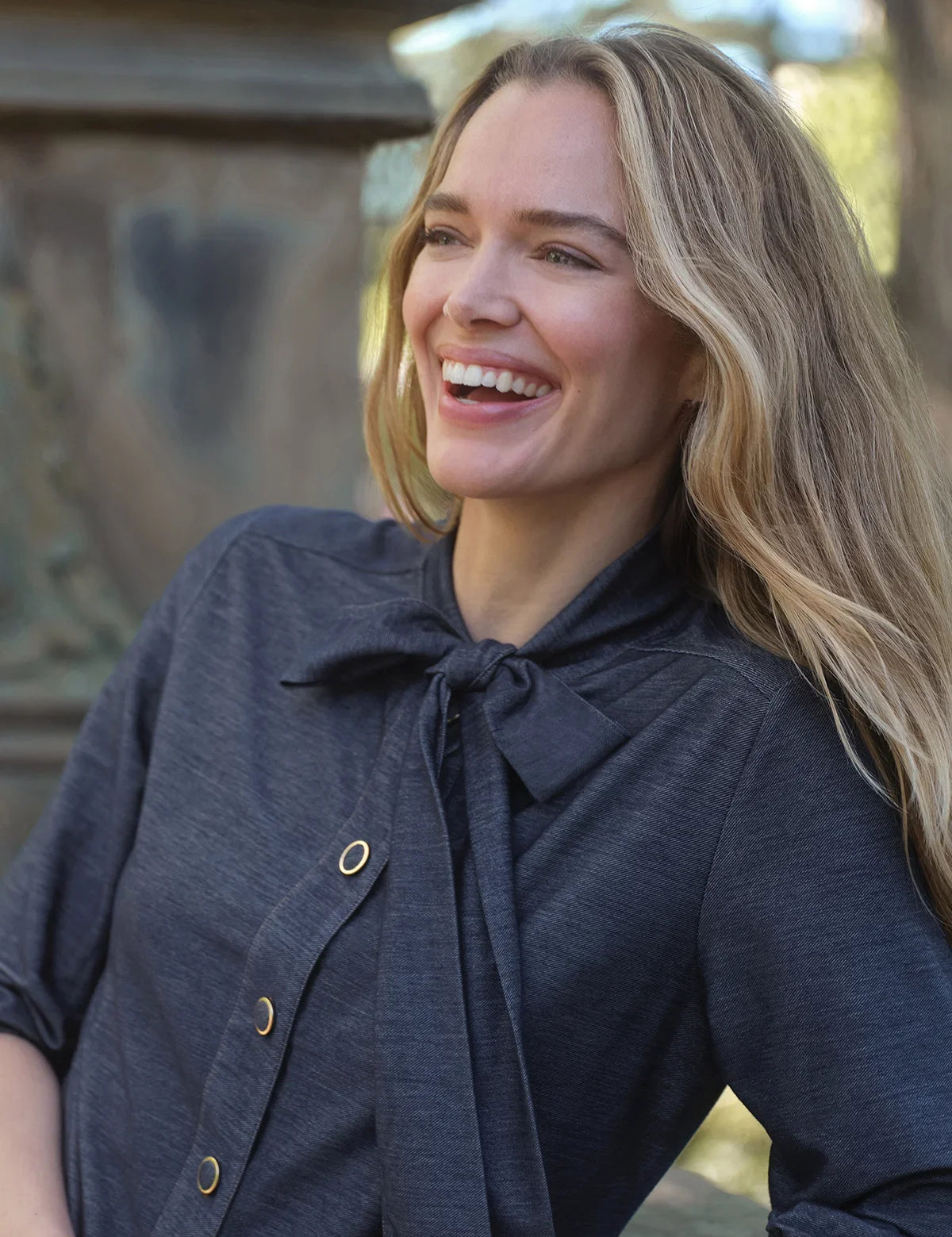 A woman with long blonde hair smiling outdoors, wearing a dark charcoal gray linen blouse with a bow tie neckline and gold-tone buttons.