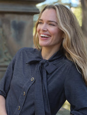 A woman with long blonde hair smiling outdoors, wearing a dark charcoal gray linen blouse with a bow tie neckline and gold-tone buttons.