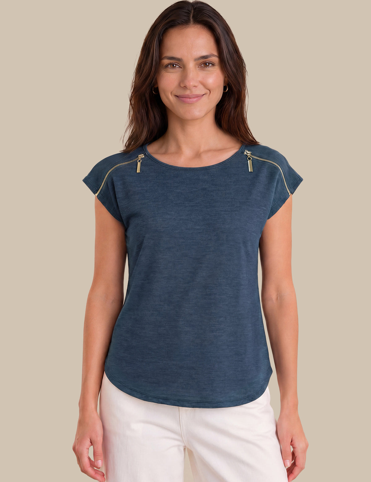 A woman wearing a slate blue short-sleeve top with gold zipper details on the shoulders, paired with white pants, set against a solid beige background.