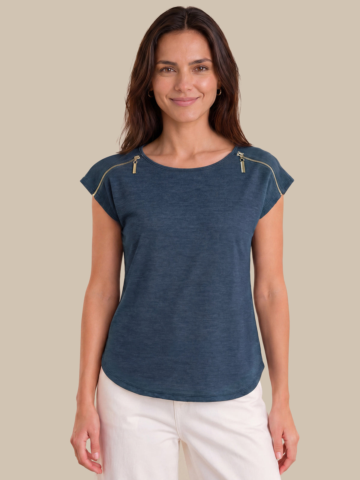 A woman wearing a slate blue short-sleeve top with gold zipper details on the shoulders, paired with white pants, set against a solid beige background.