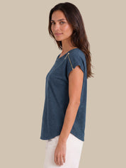 A woman wearing a slate blue short-sleeve top with a silver zipper detail on the shoulder, paired with white pants, shown in profile against a solid beige background.