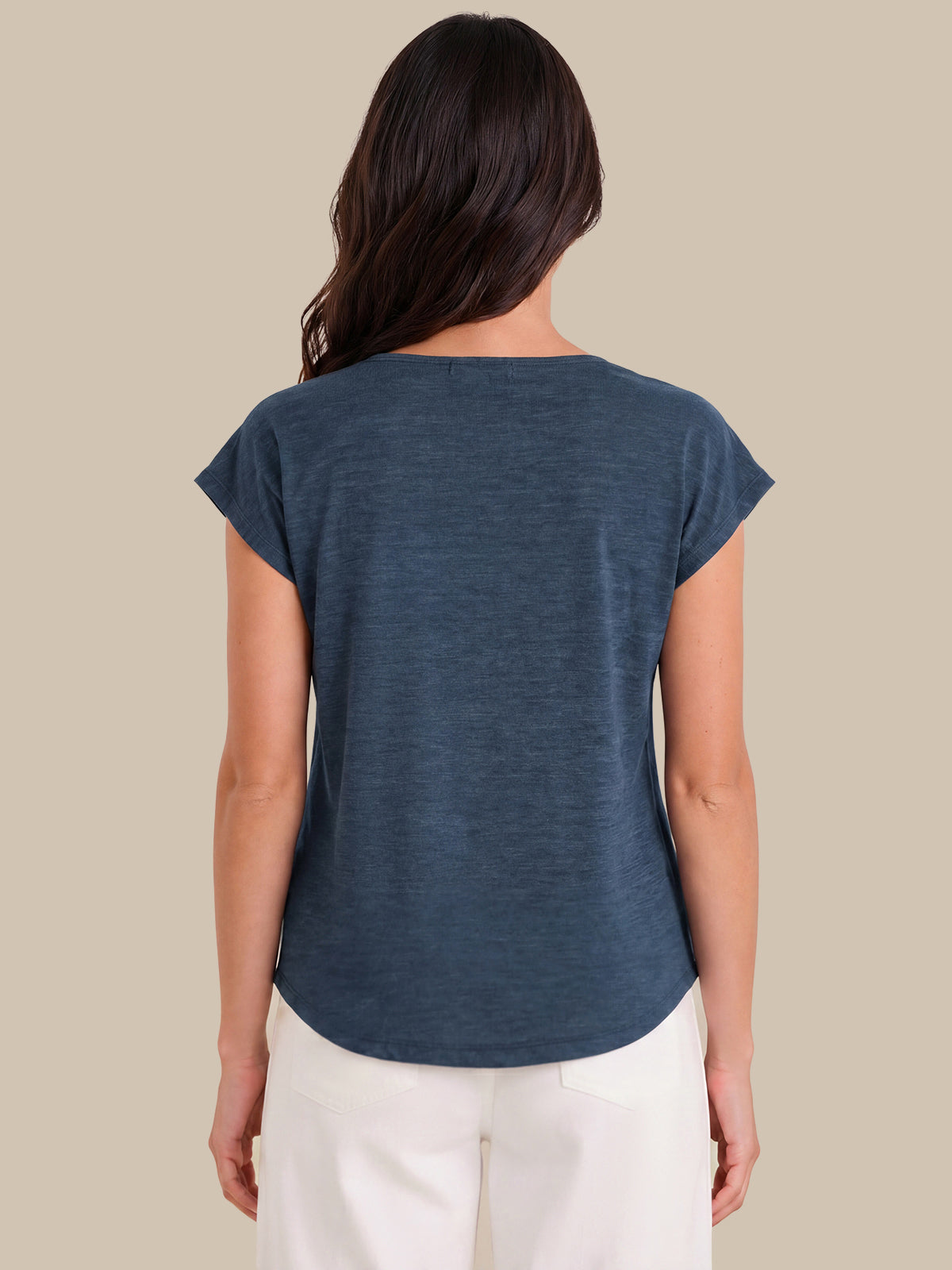 A woman viewed from the back wearing a slate blue short-sleeve top and white pants, set against a solid beige background.