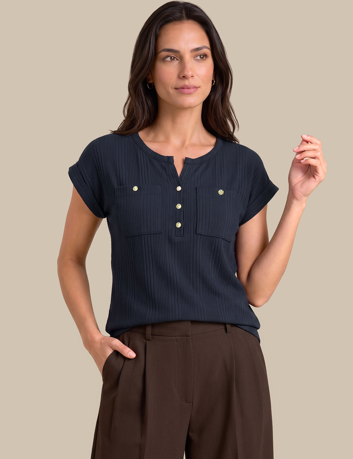 A woman modeling a dark navy blue short-sleeve top with a buttoned henley neckline and two chest pockets, paired with brown trousers, against a solid beige background.