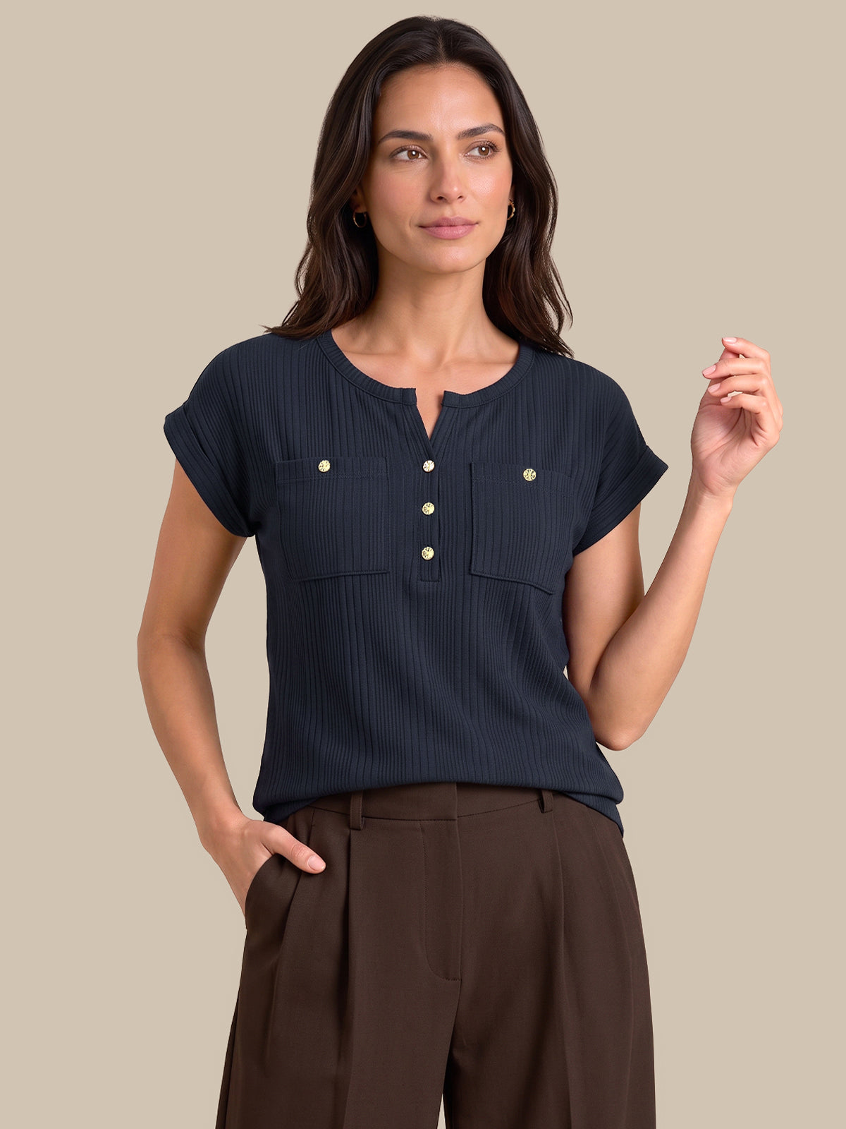A woman modeling a dark navy blue short-sleeve top with a buttoned henley neckline and two chest pockets, paired with brown trousers, against a solid beige background.