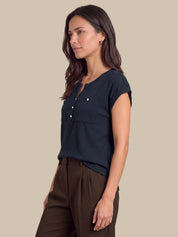 A woman wearing a dark navy ribbed short-sleeve henley top with gold buttons and brown trousers, shown in a side profile against a solid beige background.