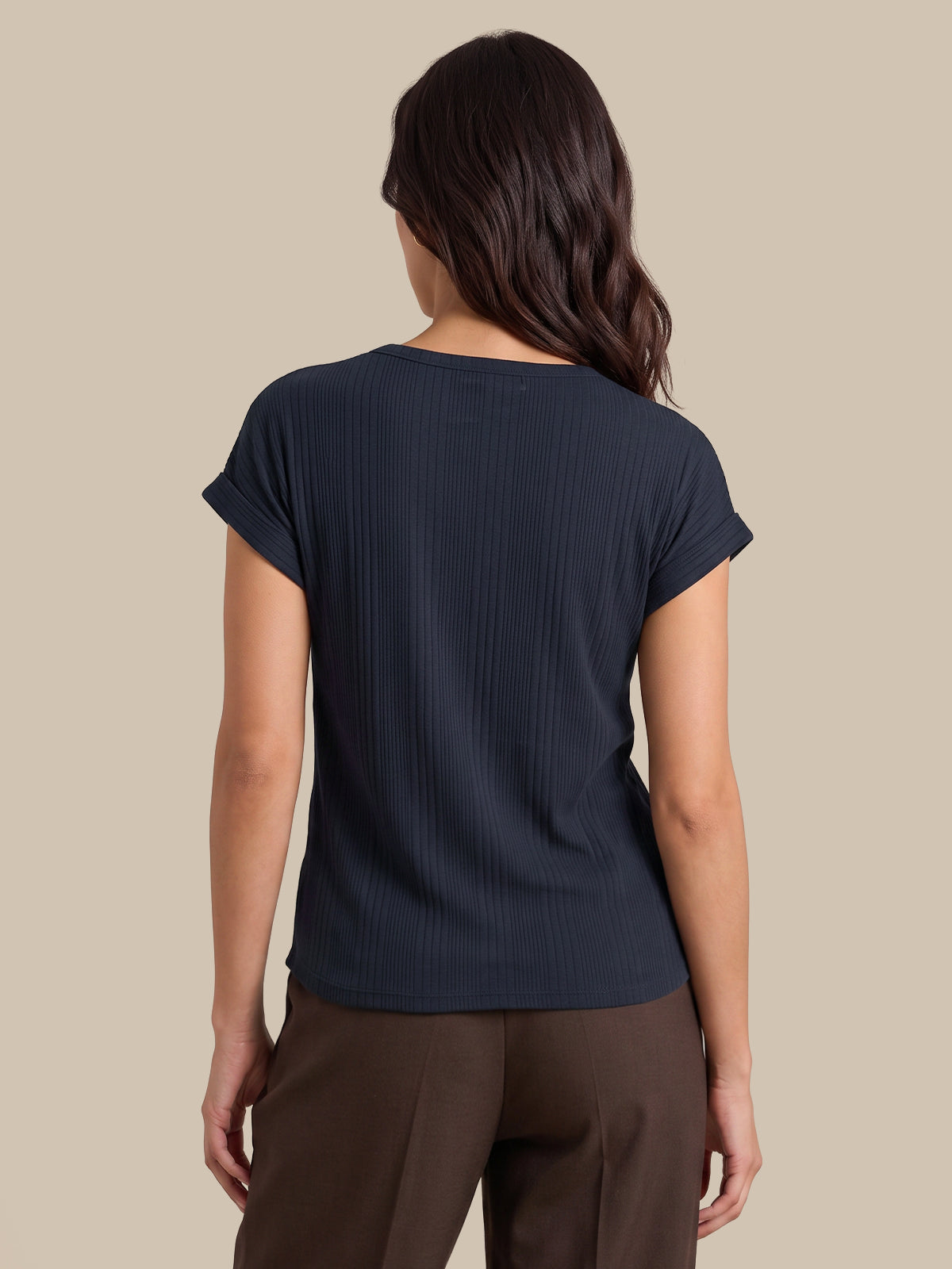 A woman shown from the back wearing a dark navy ribbed short-sleeve top and brown pants, set against a solid beige background.